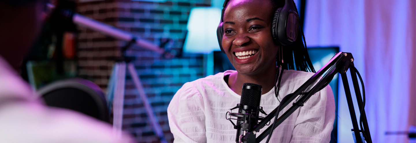 A smiling woman wearing headphones speaking into a studio microphone.