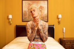 A muscular, tattooed man posing in front of a bed in a music video.