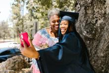 Photo of a young woman wearing a graduation gown and cap taking a selfie with her mother.
