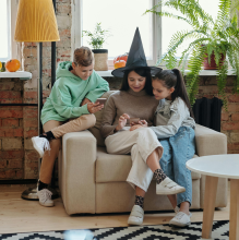 A woman and two children viewing something on the woman's phone. The woman is wearing a pointy witches hat.